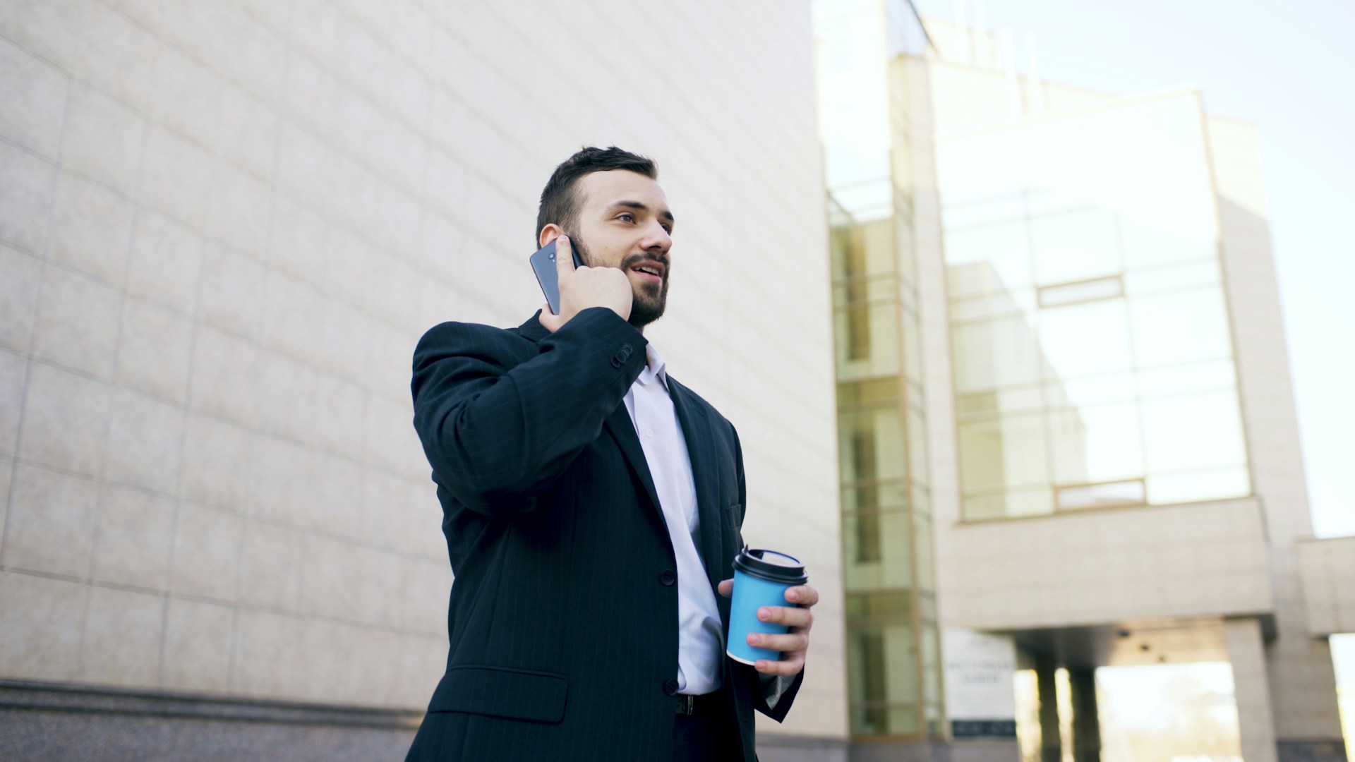 A young businessman in a suit talks on his smartphone while holding a coffee cup near a modern office building.